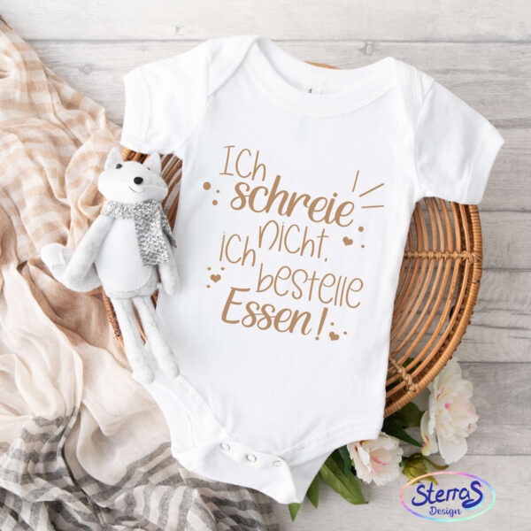 Weißer Babybody mit schwarzem Aufdruck des Designs, platziert auf einem runden Holzteller. Links daneben ein Stofftier (Pilz-Fuchs), dekoriert mit einem Schal und einigen kleinen Blumen.