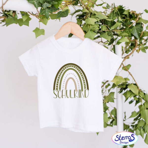 Weißes T-Shirt mit dem Regenbogen in Grüntönen und dem Schriftzug „Schulkind“. Das T-Shirt hängt dekorativ in einem Efeustrang – ein sommerliches, natürliches Setting.