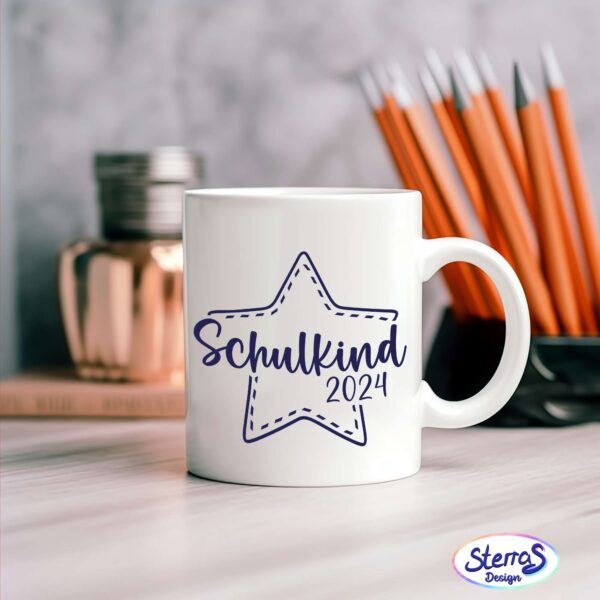 Weiße Tasse mit dunkelblauem Design: Schulkind 2024 mit einem Sternenrahmen. Im Hintergrund sind orangene Stife zu sehen. Die Tasse steht auf einem hellen Tisch.