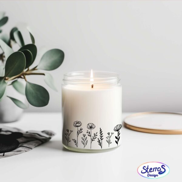 Weiße Duftkerze mit zartem Blumenwiesen-Motiv in Schwarz, dekorativ inszeniert mit Holzunterlage, Pflanze und Steinen, daneben Logo von SterrasDesign.