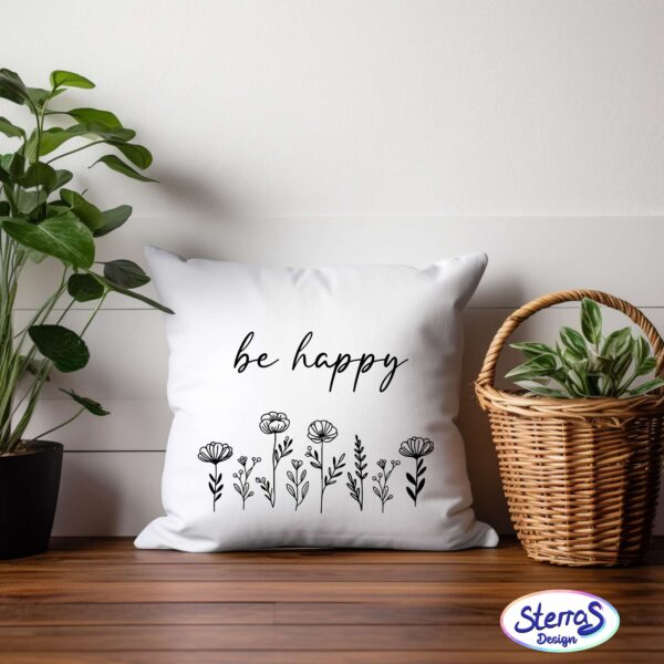 Weißes Kissen mit floralem Line-Art-Blumenmotiv und der Aufschrift „be happy“, arrangiert auf einer Holzbank mit Pflanze – ideal als Geschenk oder Dekor.