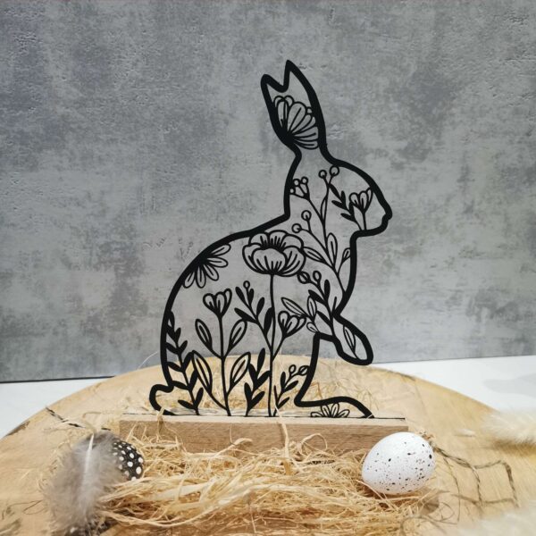 Beispielanwendung der Datei „Hase floral“: Zu sehen ist die Datei ausgeschnitten aus dicken schwarzem Karton und mithilfe von Klötzchen hingestellt. Die Deko steht auf einem Holzteller vor einem hellgrauen Hintergrund und ist dekoriert mit Dekostroh, Federn und Eiern.