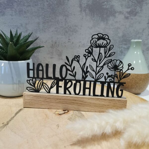„Hallo Frühling“ als ausgeschnittene schwarze Pappe in der Klötzchenvariante. Vor dem Design steht ein Klötzchen, links im Hintergrund eine Blume, rechts eine Vase. Im Vordergrund dezente Blümchen-Deko. Das Arrangement steht auf einem Holztisch mit natürlichem Licht.
