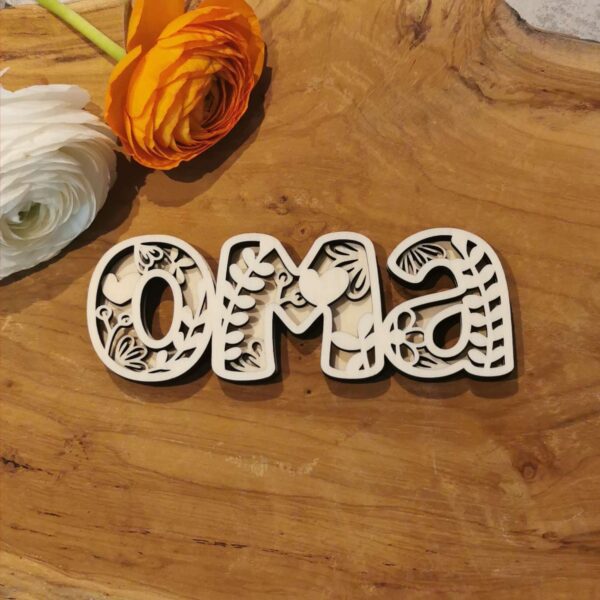 Schriftzug „Oma“ aus beigem Holz mit floralem Muster auf dunkler Rückschicht. Liegt auf Holztisch mit bunten Papierblumen – liebevoll gestaltetes DIY-Projekt.
