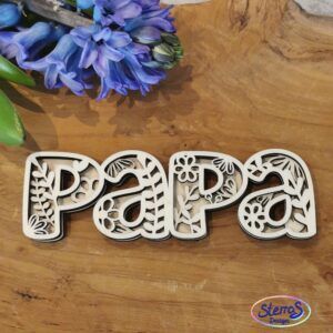Papier-Schriftzug „Papa“ mit beigem Muster vor dunkler Rücklage. Florale Elemente in beiden Schichten. Präsentiert auf hellem Tisch mit Blumen – perfekte Geschenkidee für Väter.