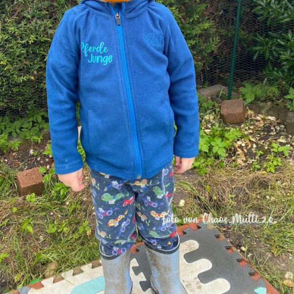 Ein Junge trägt im Garten eine blaue Jacke mit dem geplotteten Spruch „Pferdejunge“ auf der Brust. Der Kopf ist nicht im Bild. Natürliche Umgebung, authentisches Tragebeispiel.