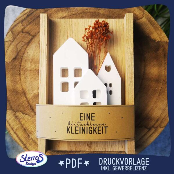 Beispielhafte Anwendung einer Druckvorlage als Banderole, angebracht um ein kleines Tablett aus Klötzchen. Auf dem Tablett liegen drei unterschiedlich große Häuser aus Raysin, darum ist die Banderole mit der Aufschrift „eine klitzekleine Kleinigkeit“ und dekoriert mit zusätzlichen schönen Trockenblumen. Auf dem Außenrahmen ist das Logo „Sterras Design“ sowie die Angabe: PDF-Druckvorlage inklusive Gewerbelizenz.