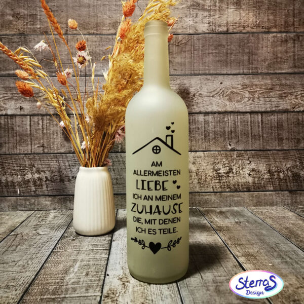 Der Spruch „Am allermeisten liebe ich an meinem Zuhause die, mit denen ich es teile.“ als schwarze Plotterdatei auf einer Flasche angebracht. Links im Bild steht eine Vase mit Trockenblumen. Untergrund und Hintergrund in warmer Holzoptik.