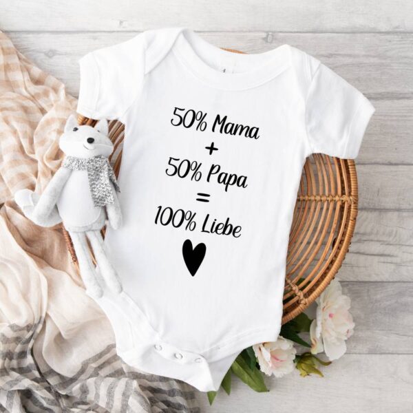 Weißer Babybody mit dem Spruch „50 % Mama + 50 % Papa = 100 % Liebe“ in Schwarz, darunter ein Herzchen. Daneben steht ein kleiner weißer Fuchs (Plüschtier), im Hintergrund ein Schal und ein zartes Blümchen.