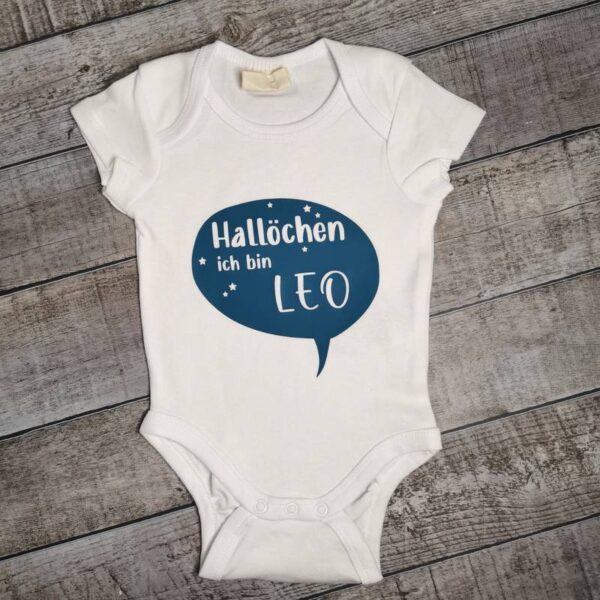 Weißer Babybody auf einem rustikalen Holzhintergrund. In türkisblauer Schrift steht: „Hallöchen, ich bin Leo“. Personalisierbares Design als süßer Willkommensgruß.