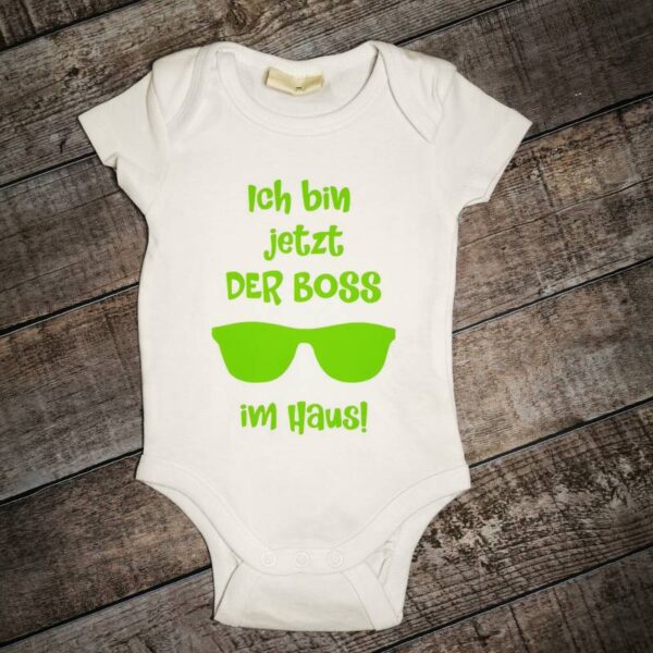 Weißer Babybody auf Holzhintergrund. In apfelgrüner Schrift steht: „Ich bin jetzt der Boss im Haus“. Unter dem Wort „Boss“ ist eine stilisierte Sonnenbrille zu sehen. Frecher Look für kleine Neuankömmlinge.
