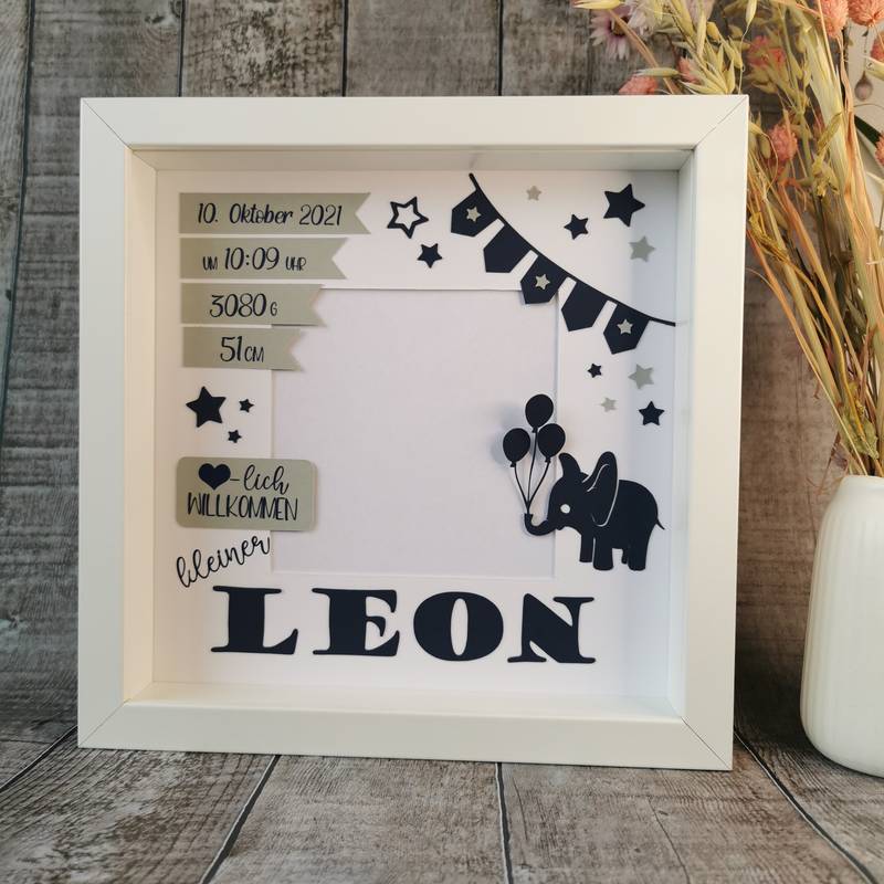 Personalisierter Geburtsrahmen in Weiß mit dem Namen LEON. Enthält Geburtsdetails wie Uhrzeit, Gewicht, Größe, Datum und Sternenmotive. Dekoriert mit Schleife und Holzoptik im Hintergrund.