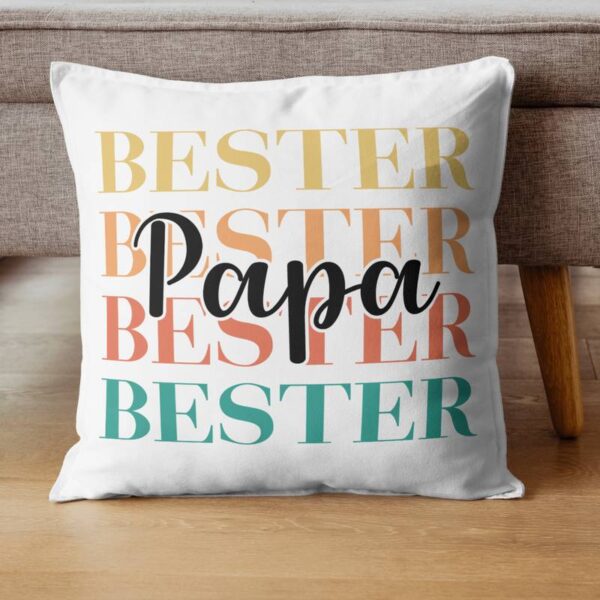 Weißes Kissen mit dem bunten Plott „Bester Papa“, dekorativ vor einer Couch platziert.