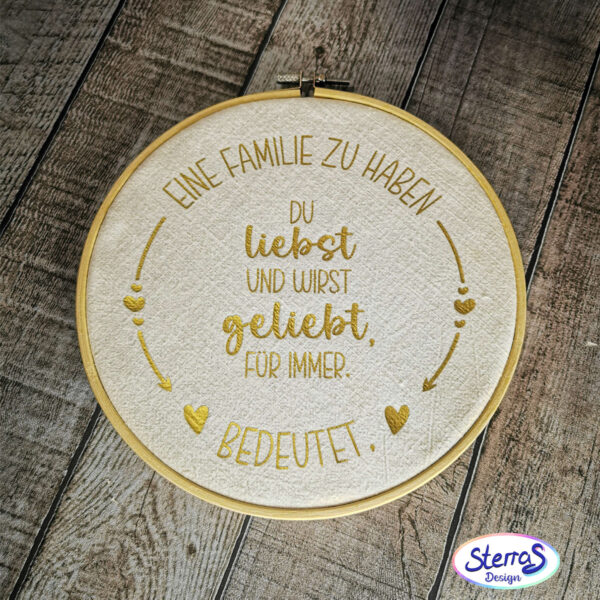 Das runde Familien-Design in Goldfolie auf hellem Stoff im Stickrahmen gepresst. Der Stickrahmen liegt flach auf einem Tisch mit Holzoptik.