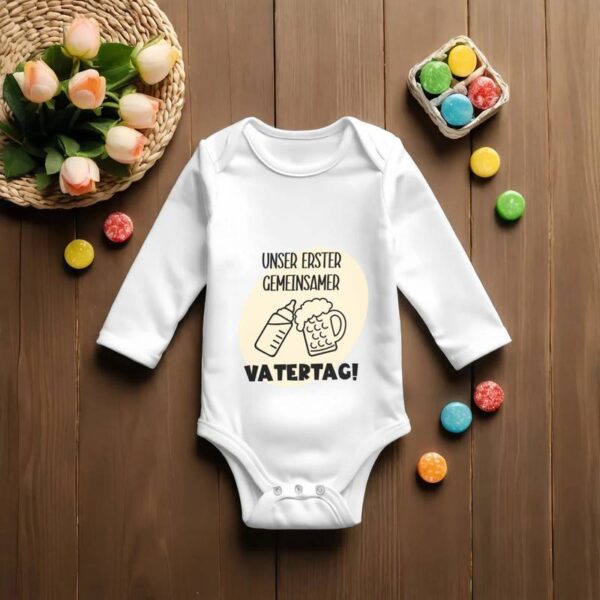 Weißer Babybody mit dem Design „Unser erster gemeinsamer Vatertag“ in schwarz und cremefarben geplottet. Um den Body herum liegen dekorativ bunte Steine, oben links sind zarte Blümchen zu sehen.
