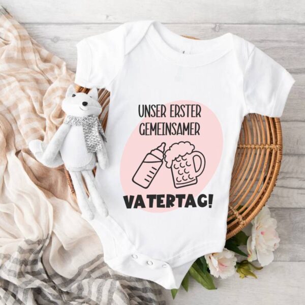 Babybody in Weiß mit dem Design in rosa und schwarz, auf einem Tisch mit hellem Holzhintergrund liegend. Daneben liegt ein Schal, ein Plüschfuchs rundet das liebevoll arrangierte Bild ab.