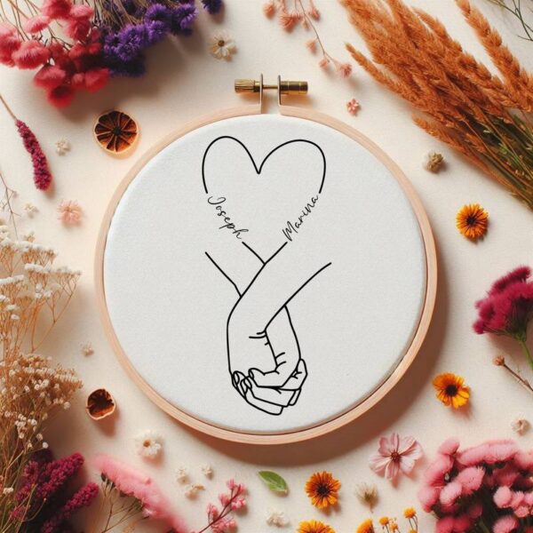 Stickrahmen mit schwarzem Hand-in-Hand-Motiv und Herzen auf weißem Stoff, mit den Namen „Joseph & Marina“, florale Tischdeko drumherum.