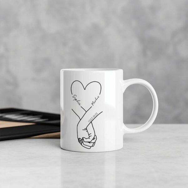 Weiße Tasse mit feinem Hand-in-Hand-Motiv im Zentrum, dekorativ auf einem hellen Holztisch platziert – schlicht und liebevoll.