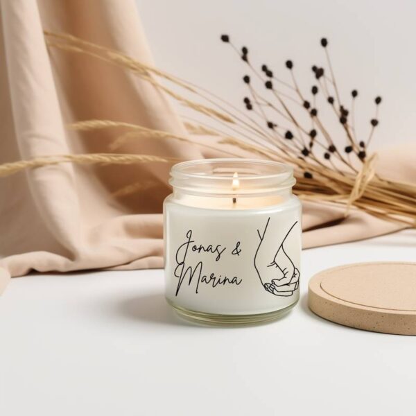 Duftkerze mit schwarzem Herz-Hand-Motiv und persönlichem Namenstext, daneben trockene Pflanzen – emotionales Geschenk für Paare.