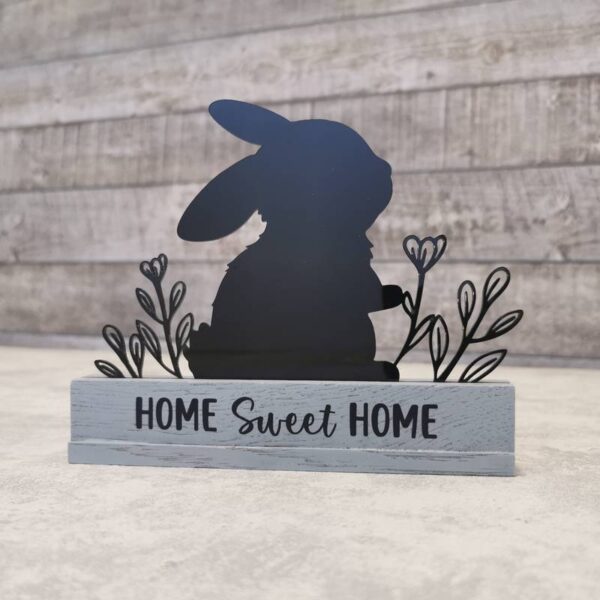 Schwarzes Acryl-Häschen eingesetzt in ein graues Klötzchen mit der Gravur „Home Sweet Home“. Der Hintergrund besteht aus Holzpaneelen, die für eine natürliche Atmosphäre sorgen.