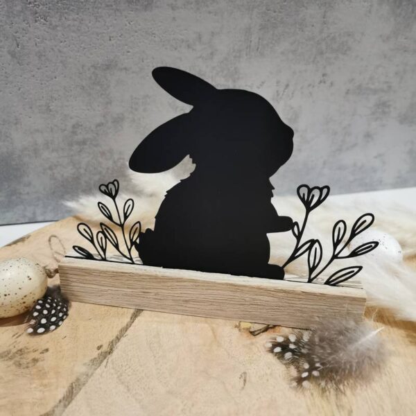 Aus schwarzem Acryl ausgeschnittenes Hasen-Klötzchen-Design mit leichtem Glanz. Feine Blumenornamente gut sichtbar. Das Dekostück steht auf einem Holztisch mit grauem Hintergrund, umgeben von Federn und dekorativen Eiern.
