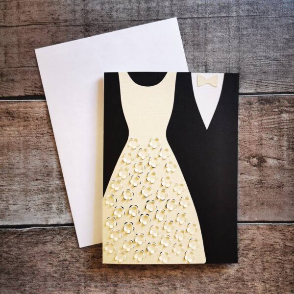 Karte mit ausgestanztem Hochzeitskleid in Weiß mit filigranem Muster und schwarzem Anzug mit Fliege. Beige Hintergrundkarte, daneben ein weißer Umschlag auf Holz. Elegante Papeterie für Hochzeiten.