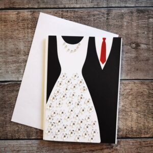 Karte mit ausgestanztem Hochzeitskleid in Weiß mit filigranem Muster und schwarzem Anzug mit roter Krawatte. Weiße Hintergrundkarte, daneben ein weißer Umschlag auf Holz. Elegante Papeterie für Hochzeiten.
