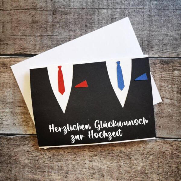 Papierkarte mit zwei stilisierten Anzügen in Schwarz mit roter und blauer Krawatte. Beschriftung: „Herzlichen Glückwunsch zur Hochzeit“. Weiße Karte mit Umschlag auf Holzhintergrund.