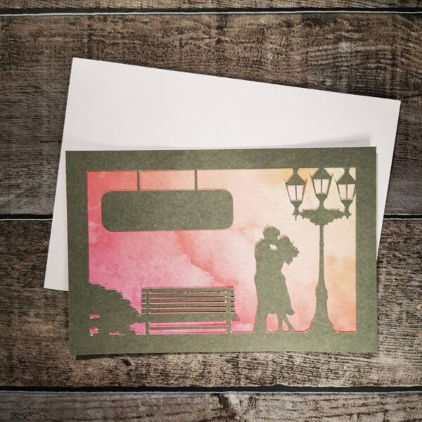 Hochzeitskarte in Silber mit einem an einen Sonnenuntergang ähnelnden Hintergrund mit wunderschönem Farbverlauf in rot und Orange. Oben links der Karte ist ein Hängeschild welches zur Personalisierung verwendet werden kann. Die Karte liegt mit einem weißem Briefumschlag auf einem Holztisch.