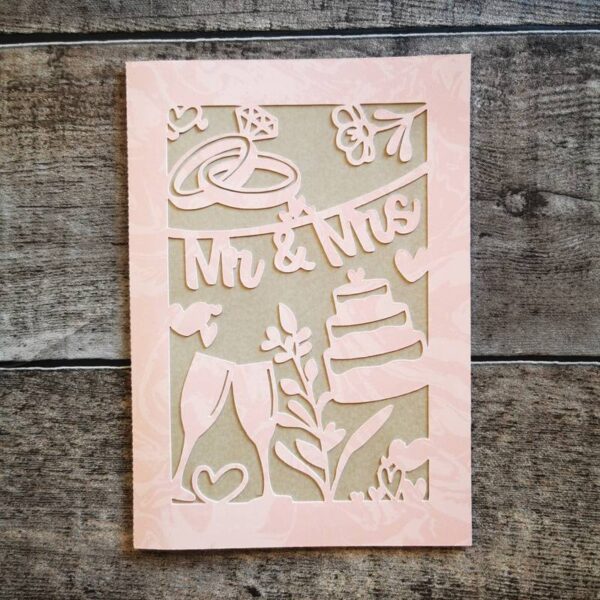 Zartrosa Papierkarte mit Hochzeitssymbolen (Torte, Gläser, Herzen, Ringe) und Schriftzug „Mr & Mrs“. Präsentiert mit weißem Umschlag auf rustikalem Holz – liebevoll gestaltet.