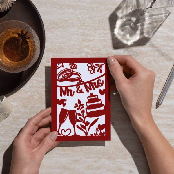Rote Karte mit filigranem Ausschnitt von Hochzeitssymbolen, gehalten von einer Hand. Auf dem Tisch: Umschlag, Tasse und Lichtspiel – liebevoll in Szene gesetzt.