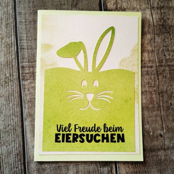 Osterkarte in Grün mit stilisiertem Hasengesicht und den Worten „Viel Freude beim EIERSUCHEN“, Karte steht auf hellem Holzuntergrund.