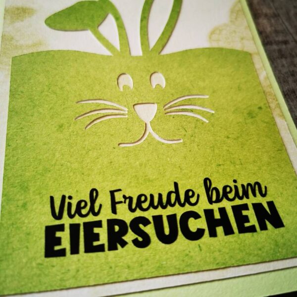 Nahaufnahme der grünen Osterkarte mit Hasengesicht und schwarzem Aufdruck „Viel Freude beim EIERSUCHEN“, feine Papierschattierungen sichtbar.