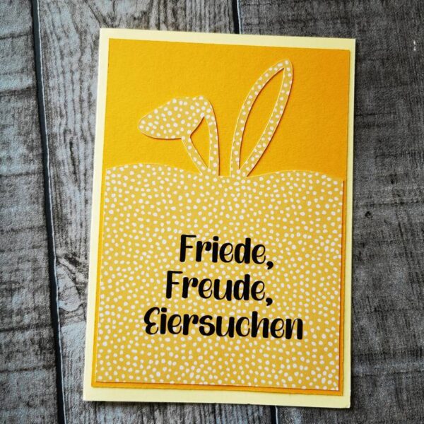 Osterkarte mit orange-gelbem Muster, Hasenohren und dem Text „Friede, Freude, Eiersuchen“ in Schwarz, Karte liegend auf einem Holztisch.