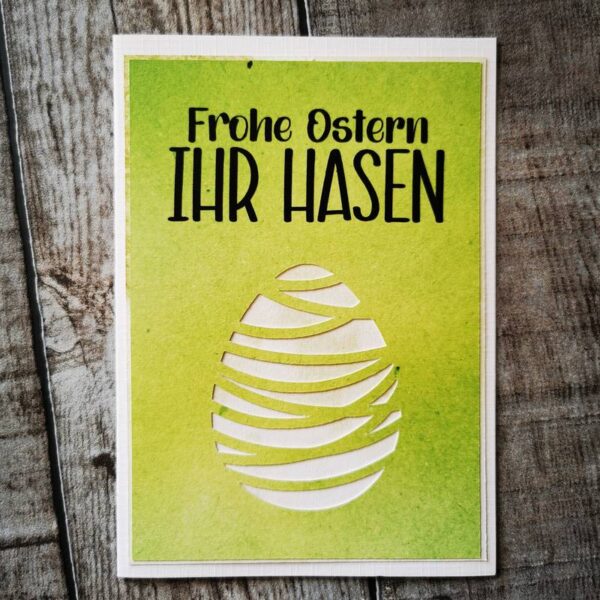 Grüne Osterkarte mit weißem Osterei und schwarzem Text „Frohe Ostern IHR HASEN“, auf hellem Holzuntergrund platziert.