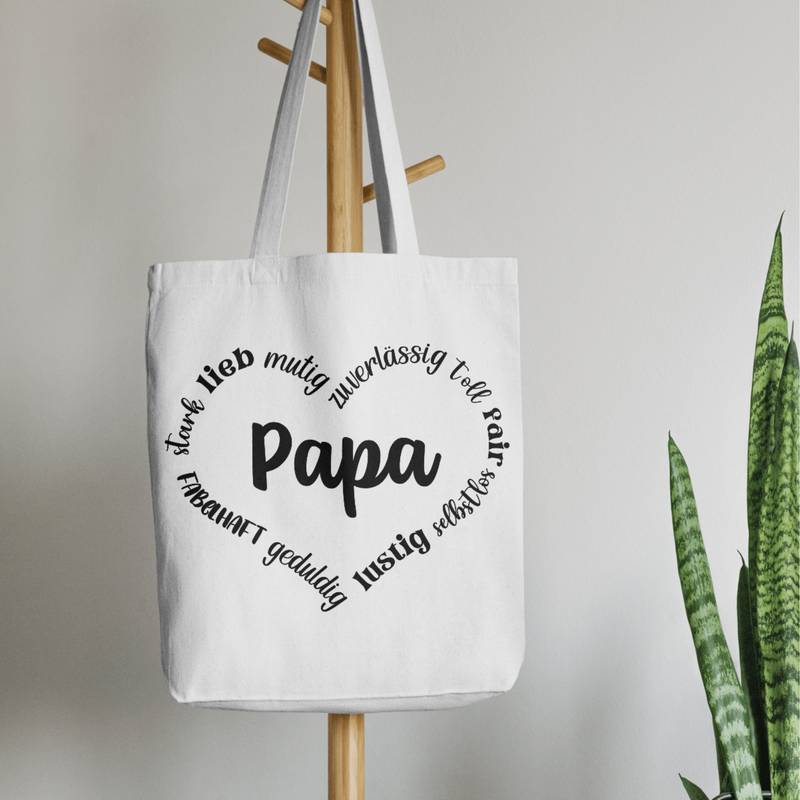 Schwarzes Design auf einem weißen Leinenbeutel. Der Beutel ist schlicht fotografiert, rechts unten ist eine zarte grüne Pflanze zu sehen, die für ein frisches, natürliches Ambiente sorgt.