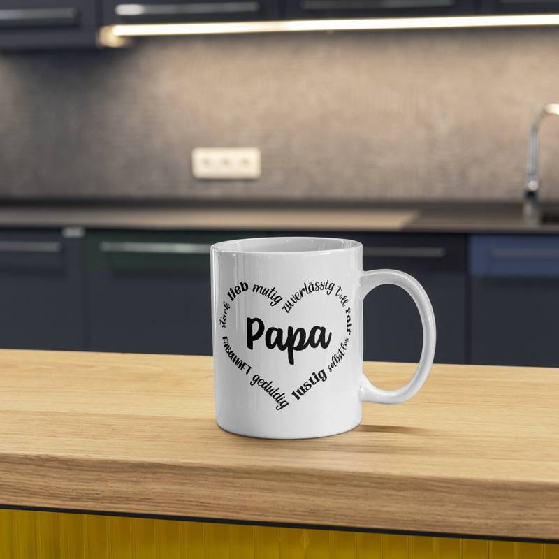 Design in Schwarz auf einer weißen Tasse. Die Tasse steht auf einem Tisch, im Hintergrund ist ein moderner Küchentresen zu sehen. Das Herz mit den Adjektiven wirkt klar und kontrastreich.