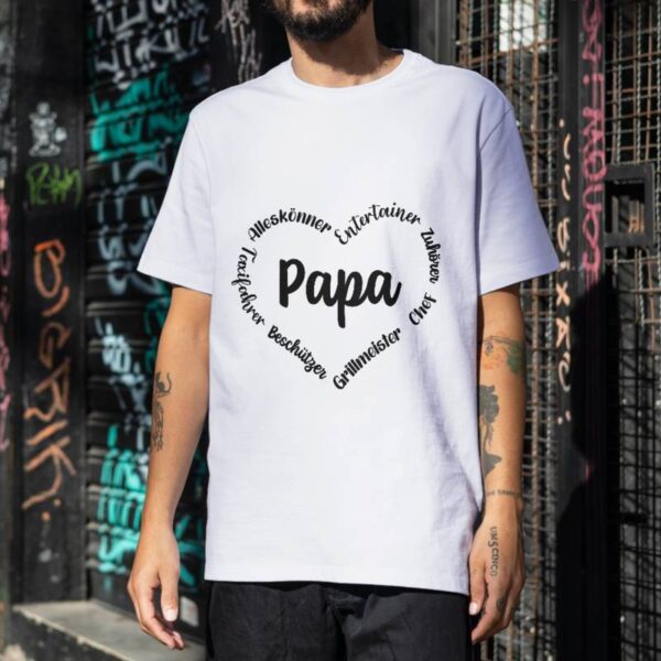 T-Shirt mit aufgedrucktem Herzmotiv, bei dem "Papa" im Mittelpunkt steht, umgeben von positiven Eigenschaften. Das Shirt wird im Alltag getragen, die Szene spielt draußen.