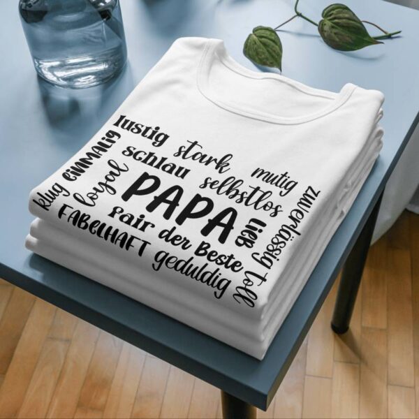 Weißes T-Shirt mit schwarzem Print des Designs „Papa + Eigenschaften“. Das Shirt liegt gefaltet auf mehreren T-Shirts an der Ecke eines Tisches. Das Design ist mittig auf der Brust aufgedruckt.