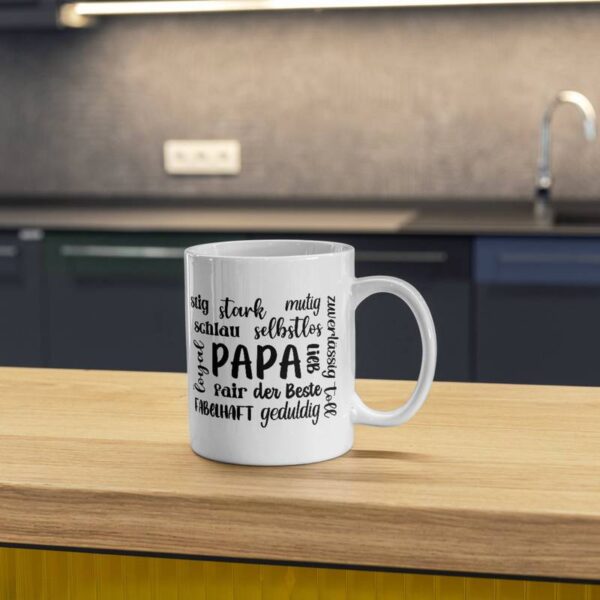 Weiße Tasse mit dem Design „Papa + Eigenschaften“ in Schwarz sublimiert. Die Tasse steht auf einem Holztisch in einer Küche, im Hintergrund sind leicht unscharf Küchentresen und -möbel zu erkennen.