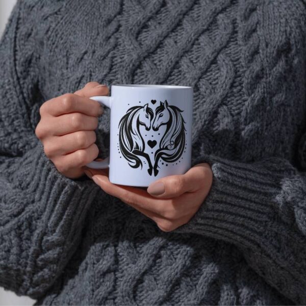 Weiße Keramiktasse mit schwarzem Motiv: zwei Pferdeköpfe die sich liebevoll anschauen. Die Tasse wird von einer Person in grauem Wollpullover gehalten – gemütliche Atmosphäre, perfektes DIY-Beispiel.