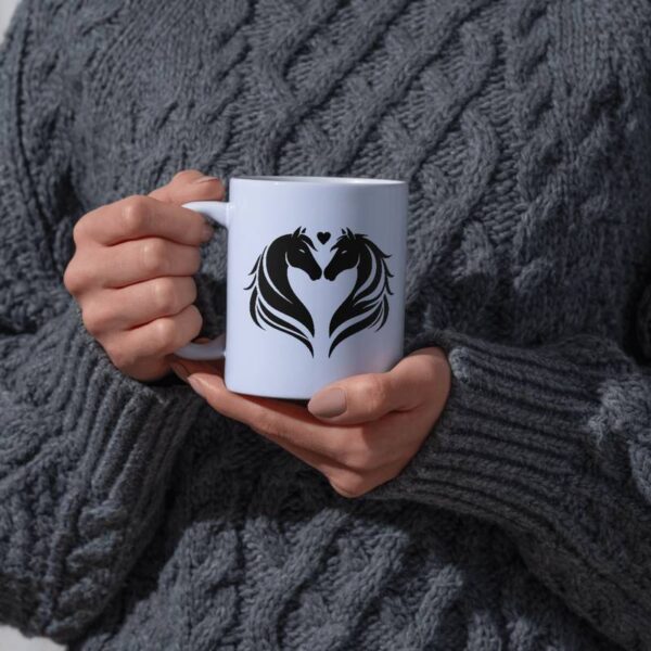 Nahaufnahme einer Tasse, die von einer Person im grauen Strickpullover gehalten wird. Auf der Tasse ist ein Design mit zwei Pferdeköpfen zu sehen, die einander zugewandt sind – Beispiel für Anwendung der Plottdatei.