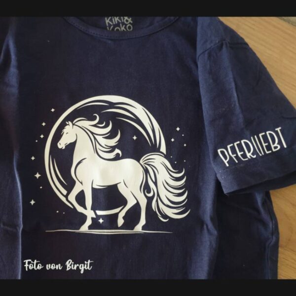 Das Design in Weiß auf einem dunkelblauen T-Shirt, in Nahaufnahme. Nur Brust und ein Teil des Ärmels sind zu sehen. Auf dem Ärmel steht zusätzlich der Schriftzug „Pferliebt“.