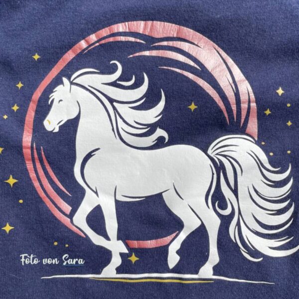 Mehrfarbige Version auf einem blauen T-Shirt, in Nahaufnahme. Das Pferd ist in Weiß, der Mond in Rosa, die Sterne in Gold. Harmonisches, fantasievolles Farbspiel.