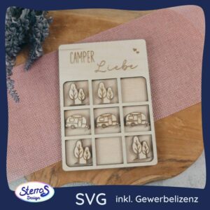 Eine Nahaufnahme eines lasergeschnittenen Holz-3-gewinnt-Spiels auf einem rustikalen Holzbrett und einer rosafarbenen Serviette. Die obere Holzplatte ist mit dem Schriftzug "Camper Liebe" und kleinen Herzen graviert. Das 3x3-Spielfeld zeigt mehrere lasergravierte Holz-Puzzleteile von kleinen Bäumen und einem Wohnwagen-Anhänger. Ein blaues Logo in der unteren linken Ecke zeigt "Sterras Design" und den Text "SVG inkl. Gewerbelizenz". Das Bild vermittelt eine handgemachte, gemütliche Camping-Atmosphäre.