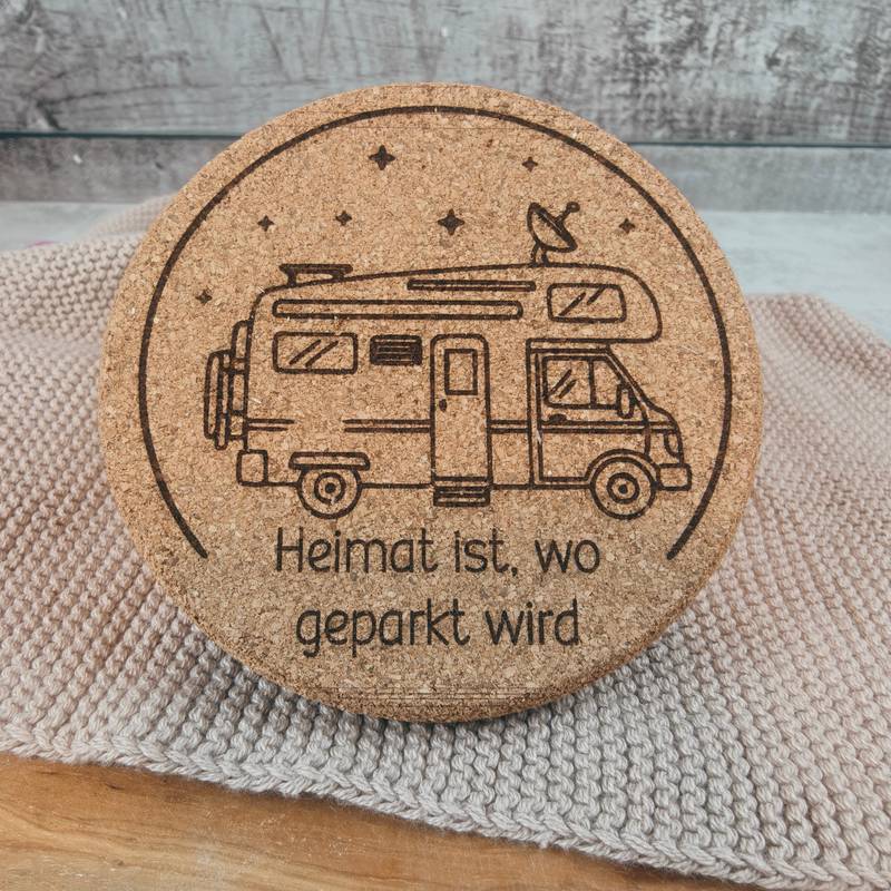 Draufsicht auf einen gravierten Untersetzer aus Kork oder dunklem Holz mit dem Motiv „Heimat ist, wo geparkt wird“, liegend auf einem hellen Stoff.